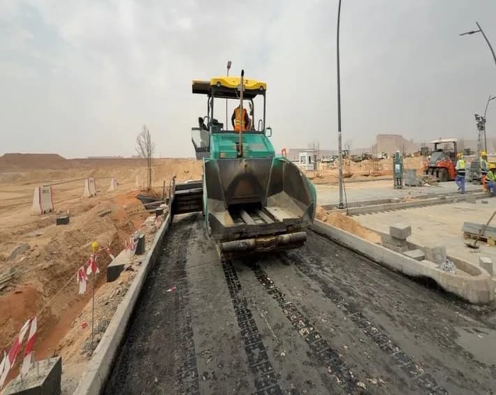 Rear view of Vogele asphalt paver on freshly laid mat with operator on deck, kerbs and taped tree pits at left, workers and truck distant at right