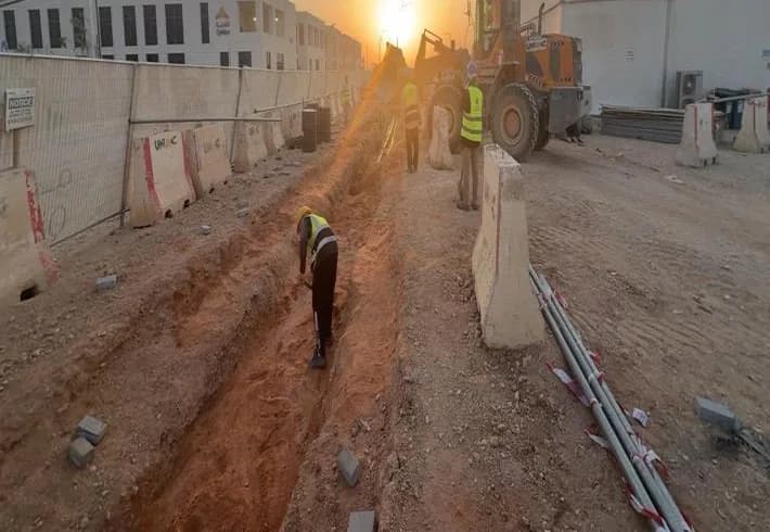 Sunset trench excavation along a fenced corridor, worker digging in trench, UNIMAC wheel loader with three workers beside it, stacked pavers, conduits and barriers, Qiddiya-logo building behind